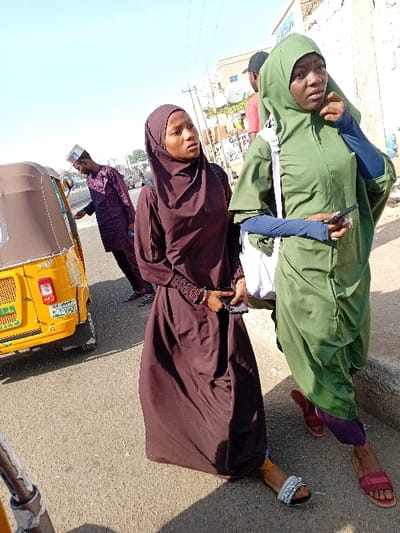 The image captures an urban street scene in Sani Mai Nagge, Nigeria, under clear, sunny daytime conditions. In the foreground, two young females stand on an asphalt road surface. The female on the left wears a dark brown full-length dress (abaya) and a matching hijab, accessorized with a dark shoulder bag. She holds a dark mobile phone near her waist and wears a silver-studded sandal on her right foot. The female on the right wears a green hijab and a loose-fitting green garment over purple trousers, with a white strap bag over her shoulder. She holds a dark mobile phone in her left hand and touches her chin with her right hand, wearing a red sandal on her left foot.
In the midground, on the left, a yellow motorized tricycle (Keke Napep) is partially visible, displaying a Nigerian license plate, MNA-193YX. A male wearing a dark purple traditional tunic (kaftan) and a patterned cap (kufi) stands partially obscured next to the tricycle. Further in the background on the right, another male is partially visible, wearing a black cap and a red shirt. The background features various buildings, overhead power lines, and a street sign with partially visible text, including "GURARA". Shadows are cast on the asphalt surface, consistent with bright sunlight.