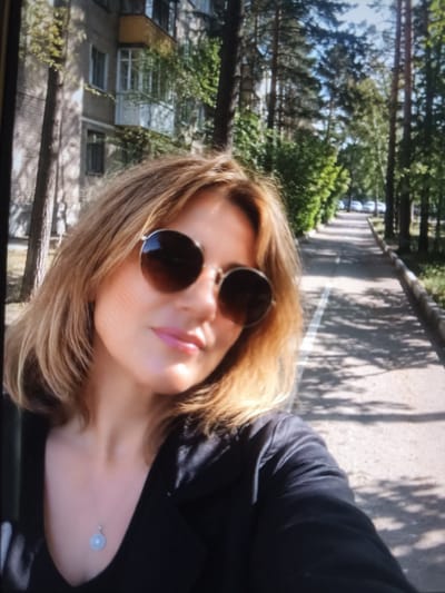 The image is a selfie taken outdoors in front of a residential building in Chekhov-2, Russia. The subject, a woman with blonde hair and sunglasses, is in the foreground. She is wearing a black top and a necklace with a blue pendant. Behind her, a paved walkway runs alongside a row of trees and a brick apartment building. The walkway is lined with white markings and dappled with sunlight and shadows. Several parked cars are visible in the background on the right side of the image.
