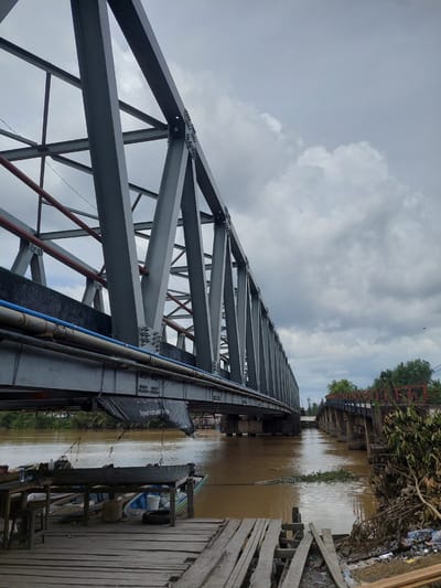 This is a vertical, low-angle shot of a gray steel truss bridge over a muddy brown river. The bridge's structure dominates the foreground, with its crisscrossing beams and bolted joints stretching towards the top of the frame. Red and blue pipes run along the side of the bridge.
The river is calm, with reflections of the sky and surrounding vegetation. On the right side of the river, a wooden walkway is visible, cluttered with various objects including boats, barrels, and what appears to be trash. Some of the boats are tied to the walkway. Lush green trees and more bridge structures are visible in the distance along the riverbanks.
The sky is overcast with dramatic, puffy clouds, suggesting it might be a cloudy day. The lighting is diffused, indicating it's likely daytime, possibly late morning or afternoon. The overall atmosphere is one of industrial infrastructure meeting a natural, albeit somewhat neglected, environment.
There is some visible text on a black tarp hanging on the side of the bridge: "PEMINDAHAN KARYA" and below it "Tingkat Kab. Bangka". This text, combined with the location context of Parit Baru, Indonesia, suggests the bridge is located in Bangka Regency, Indonesia. There are no people visible in the image.