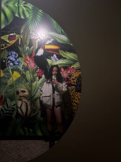 A medium shot depicts a woman posing in front of a large, semi-circular tropical mural. The mural features various elements, including a toucan, leaves, flowers, a butterfly, a meerkat, and a leopard print section at the top. The woman is holding a phone and appears to be taking a selfie. She is wearing a white jacket. The mural is situated within an arched space, with a textured wall visible on the right. The location is Amadora, Portugal.