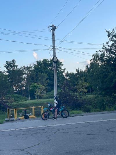 The image shows a person on a dirt bike parked on the side of a paved road. The person is wearing black shorts, a black and white patterned shirt, and black and white shoes. The dirt bike is teal and black with colorful accents on the wheels. The bike is leaning on its kickstand.
Behind the road, there is a grassy area with trees and bushes. A tall utility pole with several wires runs diagonally across the frame, stretching into the sky. The sky is clear and blue, with a few white clouds visible. A yellow gas pipe with a valve is located near the side of the road, behind the dirt bike.
The image appears to be taken outdoors during the daytime, likely in the late afternoon or early evening given the soft lighting. The setting suggests a rural or suburban area. There is no visible text in the image. The overall mood is relaxed and casual, with the subject appearing to take a break. The presence of the utility pole and the gas pipe suggests an area with infrastructure, possibly on the outskirts of a town.