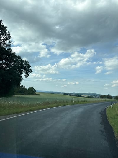The image depicts a rural roadside scene in Oberwetz, Germany, oriented vertically. The foreground features a dark asphalt road with a white line marking its edge. To the left of the road, a green grassy verge is visible, with some scattered white wildflowers. Further left, a dense, dark green tree dominates the lower part of the frame, with its branches silhouetted against the sky.
The sky is a prominent subject, featuring a mix of dramatic, heavy gray clouds and patches of bright blue sky with scattered white cumulus clouds. This suggests a dynamic weather condition, possibly a partly cloudy or transitioning weather day.
In the distance, beyond the grassy fields and a line of trees, a small village or town can be seen nestled on a hillside, with buildings and houses indicating human habitation. Rolling hills form the horizon, under the expansive sky. A single white post marker is visible along the side of the road.
The time of day appears to be daytime, likely late afternoon or early evening, given the lighting and the contrast between the clouds and the blue sky. There is no visible text in the image. The overall impression is one of a serene, yet somewhat dramatic, natural landscape with the presence of a road and a distant settlement.