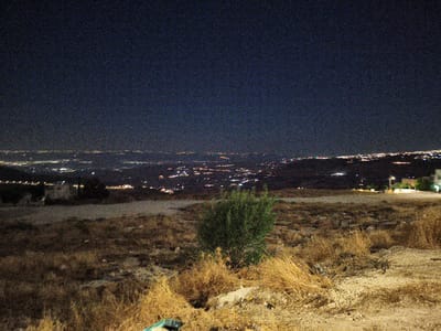 The image is a nighttime landscape photograph taken from a high vantage point overlooking a city. The sky is dark and filled with digital noise, suggesting a long exposure or low light conditions. Below the sky, a vast expanse of city lights stretches across the horizon, appearing as a dense collection of white and yellow pinpricks. The lights indicate a populated urban area.

In the foreground, the terrain is rough and arid. Dry, yellowish-brown grass and scrub cover the ground. A dense, dark green bush is visible in the middle ground. The ground is uneven, with patches of dirt and small stones. In the lower right corner of the frame, some debris is present, including what appears to be a green tarp or plastic sheeting and possibly a discarded object.

The location is identified as Umm al-Quttayn, Jordan. The scene captures a view of the city at night, likely from a hilltop or elevated area outside the city limits. There are no people or animals visible in the image. The overall impression is one of a remote, undeveloped foreground contrasting with a distant, illuminated urban center.