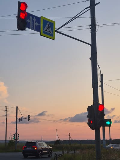 The image shows a traffic intersection at dusk in Novoe Mart'yanovo, Russia. A traffic light displays a red light with a countdown timer showing "12". Adjacent to it is a blue directional sign indicating lanes for going straight and turning left. A pedestrian crossing sign with a yellow border is also visible. Another traffic light on a pole to the right displays a red light on top and a green arrow pointing to the right. A dark SUV is stopped at the intersection, and a white car is visible in the background. Power lines are strung across the scene, and a field with tall grass and trees can be seen in the distance. The sky is a gradient of blue and orange, indicating the setting sun.