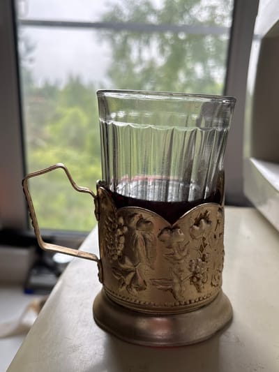 A clear glass cup, filled with a dark liquid, is held within a decorative metal holder with a handle. The metal holder has floral designs, possibly grapes and leaves. The background shows a blurred view of greenery through a window. The object sits on a light-colored surface, likely a table or countertop. The city of Ulyanovka, Russia, is not discernible from the image.