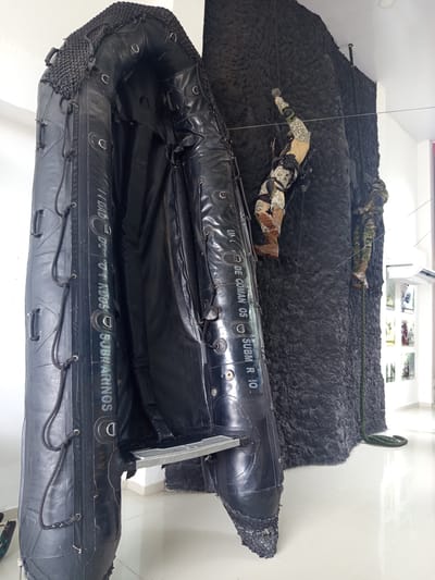 In Coveñas, Colombia, a black inflatable boat, likely a Rigid Inflatable Boat (RIB), is displayed vertically. It appears to be an exhibit, possibly at a museum or memorial. The boat is made of black reinforced fabric and features various attachment points and ropes along its sides. On the left side of the boat, vertical text reads "UNIDAD DE COMANDOS SUBMARINOS" and "SUBM R 10".
Behind the boat, a dark, textured wall simulates a rocky cliff face. Two mannequins dressed in camouflage military uniforms are positioned on this wall, appearing to climb or rappel. The mannequin on the left is higher up, with its left arm extended upwards as if reaching for a hold. The mannequin on the right is positioned lower and further back, descending with a rope. Both mannequins are wearing tactical gear, including boots, trousers, and body armor.
A green rope is visible, hanging from the top of the wall and extending down to the floor, suggesting a rappelling scenario. The floor is a light-colored, reflective surface, possibly polished concrete or tile. In the background, a white wall is visible, with a white air conditioning unit mounted high up. Further to the right, a display of framed images is mounted on the wall, hinting at the nature of the exhibit, which seems to commemorate military or naval operations. The overall scene is a staged diorama illustrating an amphibious or vertical insertion military operation.