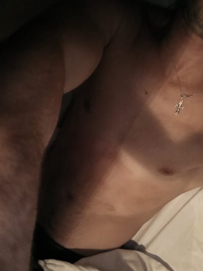 The image shows a close-up view of a person's torso, specifically the chest and upper arm, in what appears to be a dimly lit bedroom. The person is wearing a silver necklace with a cross pendant. A portion of dark fabric, possibly underwear, is visible at the bottom left of the frame, along with white bedding. The lighting creates strong shadows, highlighting the contours of the body and suggesting a horizontal orientation of the person. There is no indication of the location being Bolotnitsa, Russia, or any other specific city. The scene is devoid of any discernable action or interaction.