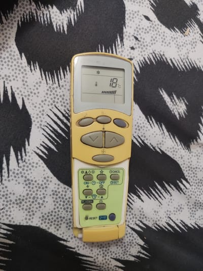 The image shows a yellow and white remote control for an air conditioning unit resting on a black and white patterned fabric. The remote control has a digital display showing "18 C" and a bar graph indicating fan speed. Below the display are various buttons for controlling the air conditioner, including temperature up/down arrows, mode selection (snowflake, sun, fan), power (on/off), and other functions like "Cancel" and "Set". The fabric has a somewhat abstract, jagged pattern. The location is described as Кипчок, Tajikistan.
