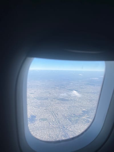 The image displays an aerial view of a densely populated urban area, observed through the elliptical window of an aircraft. The dark, metallic-grey frame of the airplane window partially obstructs the upper, left, and right portions of the view. Below, a vast grid pattern of residential and commercial buildings dominates the landscape, characterized by uniform street layouts. Several green spaces, appearing as darker, irregular patches, are integrated within the urban sprawl, with a larger, more distinct park visible towards the lower right. Small, scattered white cumulus clouds are present at a lower altitude than the aircraft, casting faint shadows onto the city below. The sky above the clouds is a clear, light blue. The scene is illuminated by daylight. No individuals or specific actions are discernible. The location depicted is the city of Martínez, Argentina.