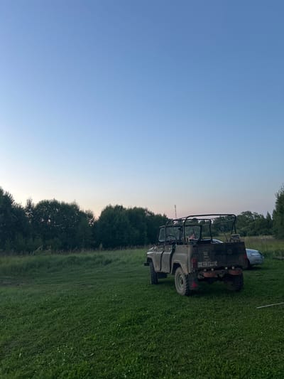 An off-road vehicle is parked on a grassy field at dusk, with a line of trees in the background. A person is visible in the driver's seat of the vehicle. A second, lighter-colored vehicle is parked to the right of the off-road vehicle, partially obscured. The sky is a clear, pale blue, transitioning to a lighter hue near the horizon. The overall scene depicts an outdoor rural setting.