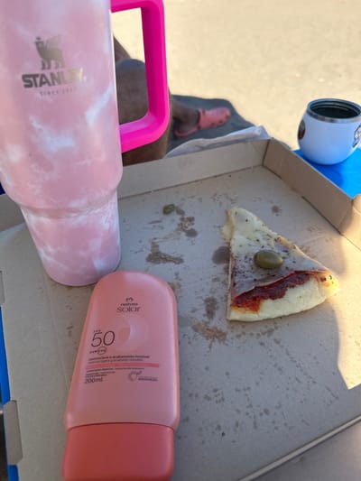 The image captures a casual outdoor scene, possibly on a sandy beach or light-colored pavement, viewed from a slightly elevated angle. In the foreground, a large pink and white marbled Stanley tumbler with a bright pink handle is prominently displayed on the left. The text "STANLEY SINCE 1913" is visible on its side. To the right of the tumbler, a pink bottle of sunscreen or sunblock is lying on its side. The label on the bottle clearly reads "natura solar FPS 50 UVA/UVB" and "textura leve e acabamento invisivel" (light texture and invisible finish) in Portuguese, along with "textura ligera y acabado invisible" in Spanish, and "200ml".
Behind these items, a partially visible cardboard pizza box contains a single, triangular slice of pizza. The pizza slice has white cheese, some red sauce, and a large green olive placed centrally on top. The pizza box itself shows several grease stains and crumbs, indicating it has been used. In the background, to the left of the pizza box, a person's leg and foot are partially visible, wearing a pink slipper or sandal, possibly a Croc. A small white and blue container, possibly a thermos or cup, is also visible near the upper left corner of the image. The lighting is bright, suggesting daytime, and creates some shadows, particularly from the objects onto the ground and within the pizza box. The overall impression is one of relaxation and leisure outdoors, perhaps enjoying a meal and drink while protected from the sun.