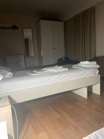A bedroom in a hotel or lodging facility in Emelyanovo (airport), Russia, is depicted. The room is sparsely furnished and appears to be a standard guest room. A white-framed bed with white linens and several folded towels is positioned in the center of the room. To the left of the bed is a small, light-colored dresser or nightstand with a metallic lamp. Behind the bed, a large white wardrobe with two doors is visible. A tall, narrow mirror hangs on the wall to the left of the wardrobe, reflecting a portion of the room, including a portion of a dark-colored bag on the floor behind the bed. A closed, light-colored curtain hangs on the right side of the room, partially obscuring the wall. The floor is covered with light-brown laminate wood. The lighting is dim, suggesting it may be evening or the curtains are drawn. There are no people visible in the image.
