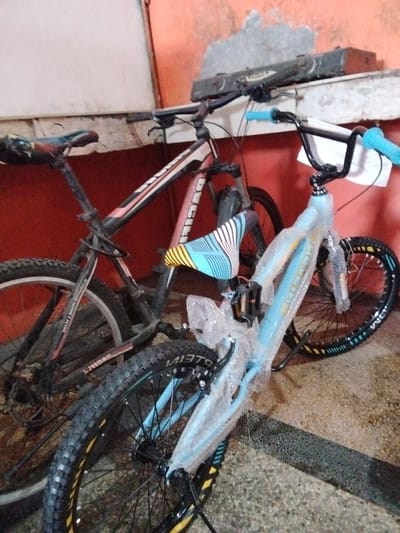 The image depicts two bicycles. The bicycle in the foreground is light blue and partially wrapped in protective plastic. It has black handlebars with bright blue grips and a black seat with blue and yellow stripes. The rear wheel of this bicycle has a black rim with yellow and light blue accents. The bicycle in the background is black and pink, with a black seat and black handlebars. Both bicycles appear to be mountain bikes. They are parked next to each other, with the foreground bicycle positioned slightly in front of and to the right of the background bicycle. The bicycles are situated on a textured, possibly concrete, floor. Behind the bicycles, there is a red wall. A shelf or ledge is visible above the bicycles, with a dark, rectangular object resting on it. The scene appears to be indoors, possibly in a storage area or shop. The overall lighting suggests it is daytime, but the environment is not well-lit. The image is taken from a low angle, looking up towards the bicycles and the wall behind them. There are no people visible in the image. The specific location in Pekanbaru, Indonesia cannot be determined from the visual information provided.