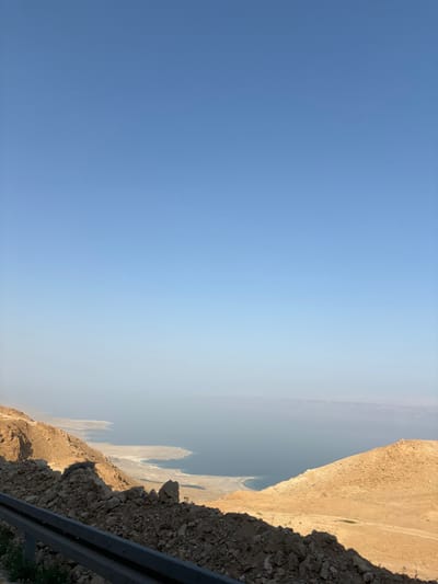 An overhead, wide shot depicts a vast desert landscape overlooking the Dead Sea under a clear, light blue sky. The top two-thirds of the image are dominated by the sky, which transitions from a deeper blue overhead to a lighter hue near the horizon. The Dead Sea, appearing a muted blue-green, occupies the mid-ground, with a visible coastline featuring white salt formations and irregular shorelines. Distant landmasses, possibly mountains, are faintly visible through a haze on the far side of the sea. The foreground shows a steep, rocky embankment composed of light brown soil, gravel, and scattered larger rocks. A dark grey metal guardrail runs horizontally across the bottom left of the frame, partially obscured by the foreground rocks. The scene is illuminated by direct sunlight, casting shadows to the left from the foreground elements. No people or specific actions are discernible. The location is consistent with the Megilot Yam HaMelakh Regional Council, Palestinian Territory.
