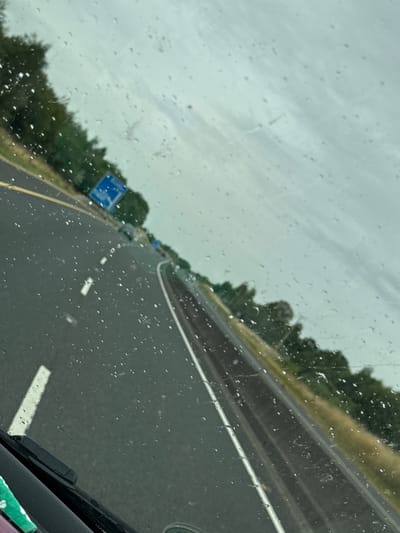 The image depicts a view from inside a vehicle on a highway, looking forward and slightly to the right. The windshield is wet with raindrops, distorting the view. The highway is paved with asphalt, with clear lane markings. On the left side of the road, there is a line of trees. On the right side, there is a grassy verge. A blue sign with white text and an arrow is visible on the left side of the road in the distance. The sky is overcast. There are no people or other animals visible in the image. The scene appears to be a typical highway environment in a rural or semi-rural setting. There is no information within the image to specifically locate it in Sheffield, Ireland.