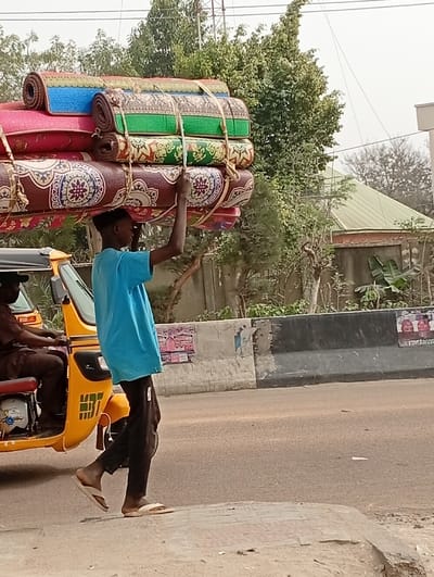 A male individual is depicted walking on a dirt and paved roadside in Darmanawa, Nigeria, balancing a substantial stack of approximately eight rolled-up, patterned mats on his head. He wears a light blue shirt, dark trousers, and light-colored flip-flops, with his hands extended upward to stabilize the load, which is secured by ropes. Immediately to his left, a yellow motorized tricycle, containing a male driver in a dark brown shirt, is either stationary or moving slowly alongside him. In the mid-ground, a concrete road barrier, painted gray and black with affixed posters, separates the pedestrian area from an asphalt road. The background consists of abundant green trees, bushes, and a building featuring a light green pitched roof.