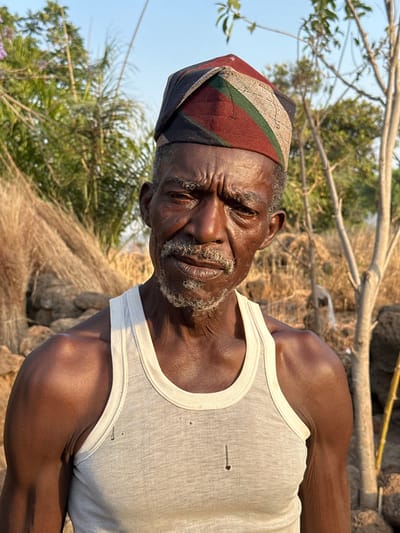 A portrait shows an elderly Black man wearing a multicolored hat and a white tank top with small holes. He gazes directly with weathered skin and graying facial hair. The background features dry grass, rocks, and greenery, possibly indicating an outdoor setting. The image is rotated, positioning the man at an angle.