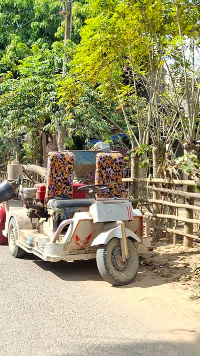 The image captures a modified, three-wheeled vehicle parked on an asphalt road. The vehicle appears to be made from salvaged parts, with a white, weathered body and colorful, patterned seat covers. It has a single front wheel and two rear wheels. A red engine or motor is visible towards the back. The vehicle is adorned with red decals of what appears to be an eagle or hawk on the front.

The backdrop suggests a rural or semi-rural setting, possibly within a village. Lush green trees and foliage line the right side of the image, with a rustic bamboo fence bordering the area behind the vehicle. A wooden utility pole with a light fixture stands prominently amidst the trees to the left. The ground is a mix of asphalt and dirt. The lighting indicates it is daytime, with shadows cast on the road, suggesting the sun is at an angle. There are no people visible in the immediate vicinity of the vehicle. The overall impression is one of a utilitarian vehicle, possibly used for transport or work, in a tropical or sub-tropical environment. The location is specified as Uleeblang, Indonesia.