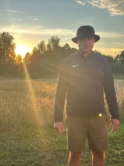 A man wearing a dark bucket hat, a dark long-sleeved shirt with a white Nike logo, and olive green shorts stands in a field of dry grass. The sun is setting behind trees in the background, casting a warm glow over the scene. The location is identified as Dubyshche, Ukraine.