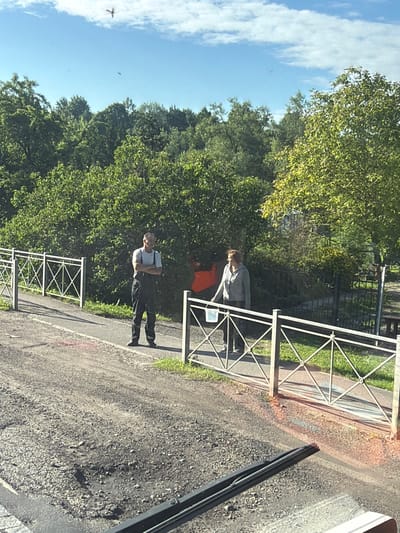 Two individuals are standing near a white metal fence and a wooded area. A man in dark overalls is standing with his arms crossed, facing to the right. A woman in a light-colored jacket and dark pants is walking to the left, holding a white bucket. The ground is gravelly with some patches of vegetation. The sky is blue with scattered clouds. In the distance, trees form a dense green backdrop. The location is identified as between rivers, Russia.
