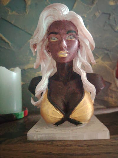 A bust of a stylized female figure is displayed on a wooden surface. The figure has dark skin, bright yellow eyes, and golden lips. Her hair is voluminous and white, styled in waves around her head. She is wearing a golden bikini top with black crisscross straps. The bust is mounted on a light-colored, rectangular base. To the left of the bust, partially visible, is a tall, cylindrical object that appears to be a pale green candle. The background features a textured wall with shades of gray and green. The overall scene is lit with warm lighting, creating highlights and shadows on the bust and surrounding objects. The location is identified as Сухарево, Russia, though no specific city landmarks are visible in the image.