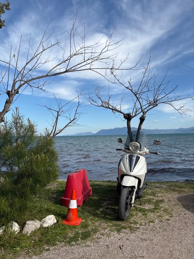 The image shows a white scooter parked on the shore of a body of water with a blue sky and white clouds above. The scooter is parked in the middle of the image facing the water. To the left of the scooter is a red and white traffic cone, and to the right of the scooter is a red plastic barricade. There are trees in the background and the water is slightly choppy. The image was taken in Vasiliki, Greece, at latitude 38.905 and longitude 22.599. It is a clear, sunny day and the water is calm. The environment is a relaxed, outdoor setting with a focus on the scooter and its surroundings.