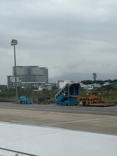 The image presents an airport tarmac scene in Hòa Cường Ward, Da Nang, Vietnam, captured from an aircraft perspective on an overcast day. In the immediate foreground, a portion of a white aircraft wing with a yellow "NO STEP" marking is visible. On the grey asphalt tarmac, a blue pushback tug with an attached trailer carrying a dark rectangular container is stationary. To its right, a blue mobile passenger stair unit is positioned, with its adjustable stairs extended upwards. Further right, a yellow cargo loader with a flatbed platform is also parked.

In the midground, a tall grey metal floodlight tower with multiple light fixtures stands erect. The background displays an urban environment featuring a prominent multi-story modern building characterized by numerous horizontal balcony or window lines. Surrounding this are several smaller, less distinct buildings and areas of green vegetation. Distant mountains, partially obscured by clouds and atmospheric haze, form the horizon under a uniformly grey sky. No people or active interactions are visible.