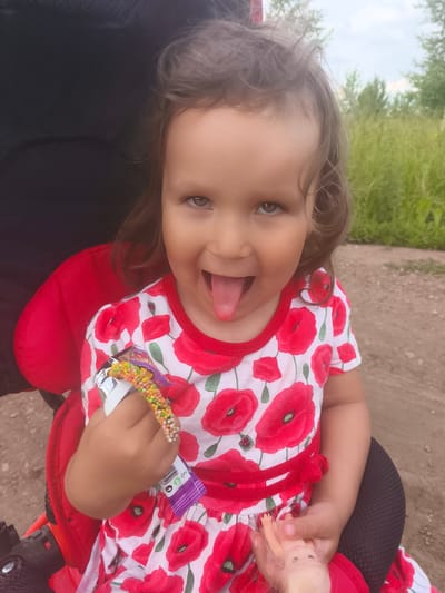 A close-up, slightly angled shot shows a young toddler girl sitting in a red stroller. She has brown hair and is sticking her tongue out at the camera. She is wearing a white dress with a pattern of red poppies. In her right hand, she holds a colorful candy stick and in her left hand, a small doll. The background suggests an outdoor setting with green foliage and a dirt path. The sky is visible, indicating it is daytime, likely with overcast weather. The overall mood is playful and cheerful due to the child's expression. There is no visible text in the image.