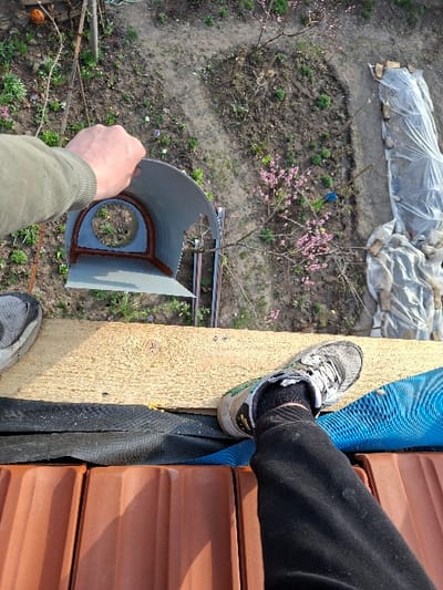An overhead view captures a scene in Krupnik, Bulgaria, from an elevated position, likely a rooftop or scaffolding, overlooking a residential garden. In the foreground, a person's lower legs, feet, and left hand are visible. The left foot, partially visible on the far left, wears a grey and white athletic shoe. The right foot, prominently displayed on the right, wears a grey, white, and black athletic shoe with white laces, a black sock, and is attached to a leg in black trousers. Both feet rest on a light brown wooden plank. Beneath the plank, a black, textured membrane or fabric is visible, covering terracotta-colored roof tiles with a corrugated profile. A blue woven material is also present under the right foot.

The person's left hand, wearing an olive-green long-sleeved garment, holds a grey, curved object, possibly a section of a gutter or vent. This object features a circular opening lined with a reddish-brown sealant or gasket material.

The background reveals a garden area primarily composed of brown, somewhat dry soil with scattered patches of green vegetation. A small tree with pink blossoms is visible towards the center-right of the garden, near which a blue plastic container or bucket rests. On the far right of the garden, an elongated area is covered with translucent plastic sheeting, secured by rocks or debris. Metal stakes or supports protrude from the ground. On the far left, a bare tree branch hangs, from which an orange, spherical object is suspended.
