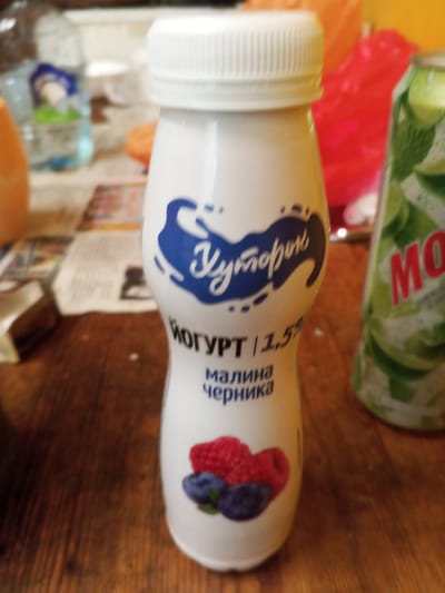 A close-up, eye-level, vertical shot shows a white plastic bottle of yogurt with a white cap, placed on a wooden surface. The bottle's label features a blue design of a milk splash or bird, with text in a script font reading "Думорок" (Dumorok). Below this, in smaller black font, it says "ЙОГУРТ | 1,5%" (YOGURT | 1.5%), followed by "малина черника" (raspberry blueberry). A small illustration of raspberries and blueberries is at the bottom of the label. To the right of the yogurt bottle, a green and white can with red lettering, possibly "MONSTER" or similar, is partially visible. In the blurred background, there are other items, including a plastic container with a blue lid and a green element, and an orange object. The wooden surface appears to be a table. The scene suggests an indoor setting, likely a kitchen or dining area, with no explicit indications of location within Ulyanovka, Russia, beyond what might be inferred from the product's branding. There are no people or actions depicted.