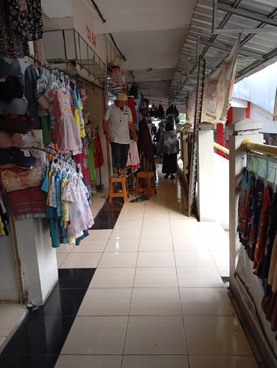 This image depicts an indoor market scene located in Pamekasan, Indonesia. The view is down a narrow, tiled walkway between vendor stalls. The floor is composed of light beige square tiles, with a strip of black tiles running along the base of the left wall. The ceiling consists of white panels and corrugated metal roofing supported by metal trusses. An overhead sign displaying "D1-S4" is visible on the left side.

On the left side of the walkway, a male individual stands elevated on two wooden stools. He is dressed in a light-colored short-sleeved collared shirt, dark trousers, and a white cap. He is looking downwards and to his right, engaged with hanging apparel. Clothing racks line the left side, displaying a variety of garments including floral dresses, children's clothing (dresses, shirts, onesies), and undergarments, all hung on hangers or stacked. A pair of dark slippers is present on the floor near the stools. Partially obscured by clothing racks further down the left aisle, another person in dark clothing is visible.

The right side of the walkway also features clothing racks displaying various garments. Further down this aisle, another individual in dark clothing and a head covering is walking away from the camera. Banners or flags are suspended from the ceiling structure on the right.