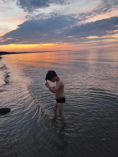 A young boy stands in shallow water near the shore of the Baltic Sea at sunset. He is wearing dark swim trunks and his head is bowed as he runs his hand through his wet hair. The sky is filled with dramatic clouds painted in hues of orange, yellow, and gray, reflecting on the calm water. The coastline, a thin strip of land with trees, is visible in the distance. The scene takes place in Apšuciems, Latvia.
