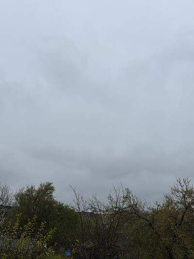 The image shows a cloudy sky above a street in Rosen, Bulgaria, as suggested by the surroundings. In the foreground and on the right side of the frame, trees and branches are visible.
