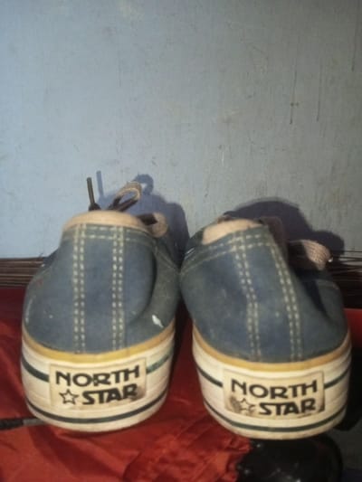 Two well-worn, blue canvas North Star sneakers are positioned side-by-side, facing away from the viewer. The sneakers feature light-colored laces and white rubber soles with a distinctive yellow stripe. The "North Star" logo is prominently displayed on the heel of each shoe, framed by black lines on the sole. The backdrop consists of a muted blue wall and a reddish-brown surface. The scene is dimly lit, casting shadows behind the sneakers.