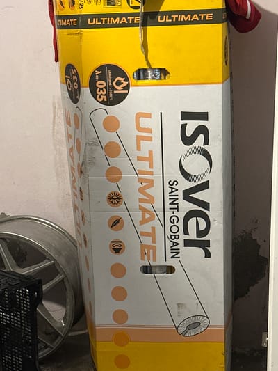 The image shows a cardboard box, likely for insulation material, leaning against a textured, light-colored wall. The box has prominent yellow and white sections with text and graphics. The text "ULTIMATE" appears multiple times in orange on a black band at the top of the box. A large logo on the side reads "ISOVER" in black text, with "SAINT-GOBAIN" below it. Various symbols, including a snowflake, a leaf, and a flame, are printed on the box.
To the left of the box is a silver-colored object with a spoked design, resembling a car wheel rim. Next to that is a black plastic crate with a grid pattern. The floor is not clearly visible.
The scene appears to be indoors, possibly in a storage area or garage, given the stacked materials and the wall texture. There are no people visible in the image. The lighting suggests an indoor environment with artificial light. The context of Ludwigshafen am Rhein, Germany, would imply this is a scene within a property located in that city.