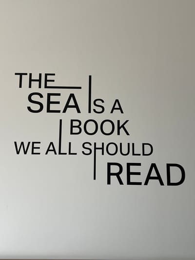 The media file is a photograph of text printed on a plain, light-colored wall. The text is arranged vertically and written in a sans-serif font, appearing to be a quote or motto. The words are black and bold, creating a strong contrast against the white background.
The text reads:
"THE SEA IS A BOOK WE ALL SHOULD READ"
The words are stacked in columns, with "THE" on the left, followed by "SEA IS A BOOK", then "WE ALL SHOULD", and finally "READ" taking up the most vertical space on the right. Vertical lines separate some of the words, acting as visual dividers.
There are no people, objects, or discernible scene elements visible in the photograph, other than the text itself. The setting is minimalist, with only the wall and the text being the focus. There are no indications of time of day, weather, or specific location cues within the image itself. The emotion conveyed is one of simplicity and emphasis on the message of the text. The context provided indicates the location is Ras Al-Khaimah, United Arab Emirates.