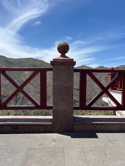 A close-up view of a red, metal fence with an X-shaped pattern, set against a backdrop of a rocky, green landscape. The fence is made of two pillars, the left one capped with a spherical stone ornament. The scene is viewed from a tiled balcony, looking down at the terrain. The sky is blue with a few white, puffy clouds. The coordinates 27.995, -15.615, place the image in the town of Tejeda, on the island of Gran Canaria, in the Canary Islands, Spain.