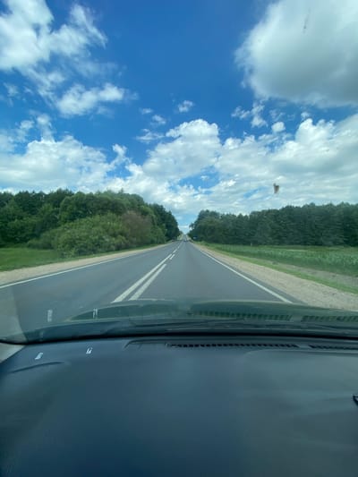 This media file captures a view from inside a vehicle traveling along a paved road bordered by lush green trees and vegetation. The road has double white lines, indicating a two-way street. The sky above is a vibrant blue, dotted with fluffy white clouds. The sun is high, suggesting it is daytime, likely late morning or afternoon, and the weather appears pleasant.
The perspective is from the driver's side, showing part of the car's interior, including the dashboard and windshield. There are some small dark specks on the windshield, possibly insects or dirt, and a blurry object that may be a bug or debris against the sky. The scene appears to be a rural setting in Belarus, given the location context of Квасоўскі сельскі Савет. The overall mood conveyed is one of a peaceful drive through a scenic, natural landscape. No people are visible, and the focus is on the road and the surrounding environment.