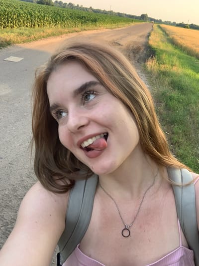 A young woman with blonde hair and green eyes is photographed from the chest up, outdoors. She is sticking her tongue out and looking to her right, with a playful expression. The woman is wearing a pink top and a silver necklace with a circular pendant. A gray backpack is visible on her shoulders. The background consists of a dirt road, a field of green crops, and a field of golden crops. The setting appears to be rural, possibly in or near the village of Derevky, Ukraine, given the presence of agricultural fields and a dirt road. The lighting suggests it is late afternoon or early evening, with the sun casting a warm glow on the scene.