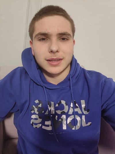 The image presents a close-up, chest-up view of a young male subject situated indoors in Kortrijk, Belgium. The individual has short, light brown hair, exhibits light stubble on his chin and upper lip, and possesses a light complexion with dark eyes. He maintains a direct gaze towards the camera with a neutral expression, his lips slightly parted. He is wearing a royal blue hooded sweatshirt, or hoodie, with visible drawstrings. The front of the hoodie displays the brand name "JACK & JONES" printed in a grey, black, and white camouflage pattern. The background consists of a plain, light-colored wall, and a portion of a light-toned upholstered chair or sofa is visible on either side of the subject.