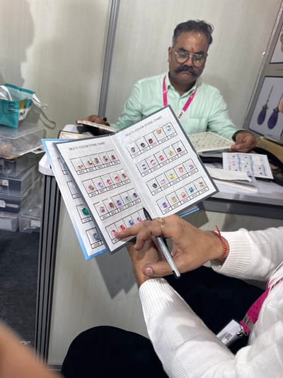 The scene depicts an interaction at what appears to be an exhibition booth or office within Jaipur Municipal Corporation, India. In the foreground, a person, identifiable by their hands, is holding open a "MULTI-COLOR STONE CHART" book. The right hand, adorned with a red thread on the wrist and a gold ring on the index finger, holds a black and silver pen and points to a specific stone sample labeled "BX17," a red-pinkish faceted stone, on the chart. This person wears a white knitted long-sleeved sweater and a pink and white lanyard with an ID badge.
In the mid-ground, seated at a light-colored table, is a middle-aged man with a dark mustache, round spectacles, and a red tilak on his forehead. He wears a light green shirt and a pink lanyard. He is looking down at documents spread on the table in front of him, with his hands resting on them.
The "MULTI-COLOR STONE CHART" displayed prominently contains numerous small, individually packaged gemstone samples of various colors and cuts, each numerically coded (e.g., BX01, BX21, UV09). The table also holds additional documents. To the left, a stack of clear plastic containers filled with small items and a teal and white bag are visible. To the right, behind the seated man, part of a display showing larger jewelry pieces is present. The background consists of a plain light-colored partition wall.