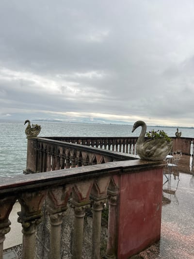 A high-angle, eye-level shot reveals a stone balcony adorned with a swan statue on an overcast day, with the city of Desenzano del Garda, Italy, in the distance. The weathered stone balcony has a rusty red section and is built on top of pillars. The swan statue sits atop a rectangular section of the balcony and is planted with greenery. Below the statue is a series of dark triangle designs that sit atop cylindrical pillars with triangular designs. In the background is the lake, with the outline of the shoreline visible in the distance. Above that is the gray, overcast sky. Visible in the very top portion of the photo is a small stone swan on another balcony area, as well as white metal lawn furniture and a wet cobblestone ground.