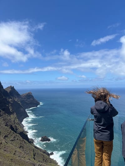 A person stands at the edge of a glass platform, gazing down at a rocky coastline and turbulent sea in La Aldea de San Nicolás, Spain. The subject, facing away from the camera, has long, windblown hair, wears a dark jacket, and tan pants. The glass platform provides a clear view of the cliffs, crashing waves, and a small, rocky islet. Above, a blue sky is dotted with scattered clouds.