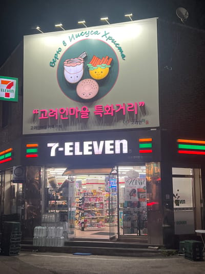 A nighttime exterior shot of a 7-Eleven convenience store in Gwangju, South Korea, is captured from a slightly low angle. The store's prominent illuminated sign reads "7-ELEVEN" in white text with red, green, and orange horizontal bars on either side. Above the store, a large, rectangular billboard features a circular graphic with cartoonish depictions of food items, including a cup with a pink drink, a bowl of orange sticks, and a pink donut, all with faces. Text in a stylized font is present on the billboard in both Cyrillic and Korean script. The store entrance is visible, with shelves of products displayed inside, and stacks of bottled water and green plastic crates are positioned outside. The surrounding environment is dark, suggesting the image was taken at night.