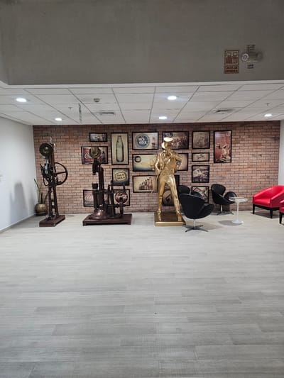 Here is a description of the image:
The image shows the interior of a commercial space, possibly a waiting area or a lobby, in La Molina, Peru. The background wall is made of red bricks adorned with framed Coca-Cola and Inca Kola advertisements. In the center stands a golden statue of a person. There are black chairs with small white tables nearby. To the right is a floor made of grey tiles. A series of vintage-style contraptions are placed along the wall. In the upper left corner, there is a section of the ceiling showing light fixtures and emergency exit signs.