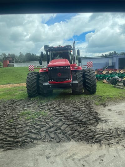 A large red tractor with "КИРОВЕЦ" written on the front is parked on a muddy surface in front of a grassy field. The tractor has massive tires with deep treads. In the background, there is a long, blue building with a white door and stairs, and several agricultural plows are lined up on the grass. Trees and a cloudy sky are visible behind the building. The scene appears to be in an agricultural or industrial area.
