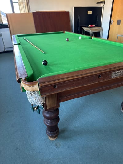 The image displays an indoor scene in Braintree, United Kingdom, featuring a large, dark wooden billiards table with a green felt playing surface. On the felt, a white cue ball, a black eight-ball, and a dark brown striped ball are scattered. A light-colored wooden cue stick with a blue tip rests on the table. The table's frame is dark wood, distinguished by carved, turned legs, and a brass-colored nameplate on its side, partially legible as 'PARKER & SONS'. A fabric net pocket, exhibiting minor fraying at its rim, is attached to the foreground corner. The floor is covered with a light blue-green carpet, displaying several small discolored spots. In the background, two large rectangular wooden panels, one light brown and one dark brown, lean against a light-colored wall. To the right, a black rectangular panel is positioned against the wall, with four light-colored tables stacked in a nested configuration in front of it. A triangular rack holding a full set of billiard balls is placed on the uppermost nested table. A window is visible on the left wall, and a light-colored wooden door with a silver handle is partially visible on the far right. No individuals are present.
