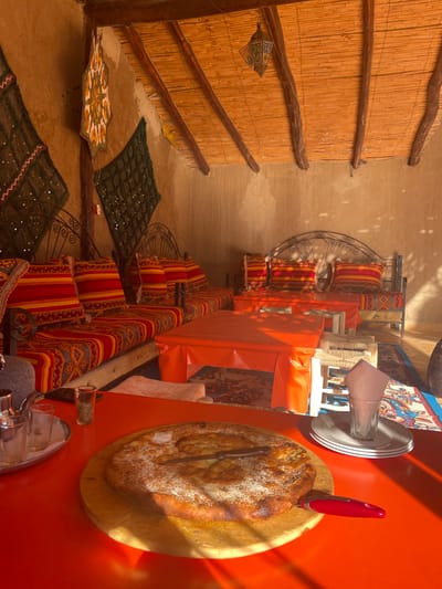 A rustic interior features low seating with patterned cushions and mats on the floor. A wall is covered in textured material with exposed wooden beams. Tables are draped in orange fabric, holding bread on a wooden board and serving utensils. Pillows are stacked on a metal frame along the wall.