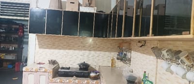 The image displays an indoor kitchen or utility area situated in Pontianak, Indonesia. The scene is devoid of occupants.
The walls are partially covered with light-beige rectangular ceramic tiles arranged horizontally. On the right, a tiled countertop holds a yellow bottle of cooking oil, a clear plastic bag, two green bottles, and a dark-colored appliance. Below this counter, a black metal rack suspends multiple white and beige mugs.
In the central area, another tiled countertop features a black two-burner gas stove with a dark pot placed on the left burner and a metal teapot to its right. Above the tiled backsplash, a row of upper cabinets or shelves with dark panels and light-colored frames are mounted. Cardboard boxes are stacked on top of these cabinets.
On the left, a multi-tiered open shelving unit contains footwear, a red bag, and various household containers. Adjacent to the lower part of this unit, two red containers rest on the floor. A white slatted structure, possibly a window or vent, is visible in the upper left section of the frame.