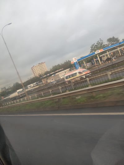 The image depicts an outdoor urban scene under an overcast sky, captured from a moving vehicle. In the immediate foreground, a dark asphalt road surface is visible, featuring a faint yellow lane demarcation line. A metal guardrail runs horizontally across the mid-ground, separating the foreground road from an elevated multi-lane highway or railway line.

On the elevated thoroughfare, vehicles are in transit, exhibiting motion blur. A white and orange passenger vehicle, possibly a train or large bus, moves from right to left. A white tanker truck is also visible, along with several smaller cars and vans. A tall, dark lamppost stands on the far left.

Beyond the elevated roadway, a busy OLA Energy gas station is prominent. It features a blue and white canopy sheltering multiple fuel pumps. Several individuals are present at the gas station, some near the pumps and others around an adjacent building structure, indicating active patronage. Behind the gas station and to its left, a multi-story, light-colored building with numerous windows rises against the horizon. Various trees and green vegetation are scattered throughout the scene. The setting is identified as Theta ward, Kenya.